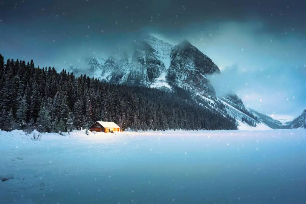 Snow falling on Lake Louise in December with a wooden cottage glowing at the lake shore in the back