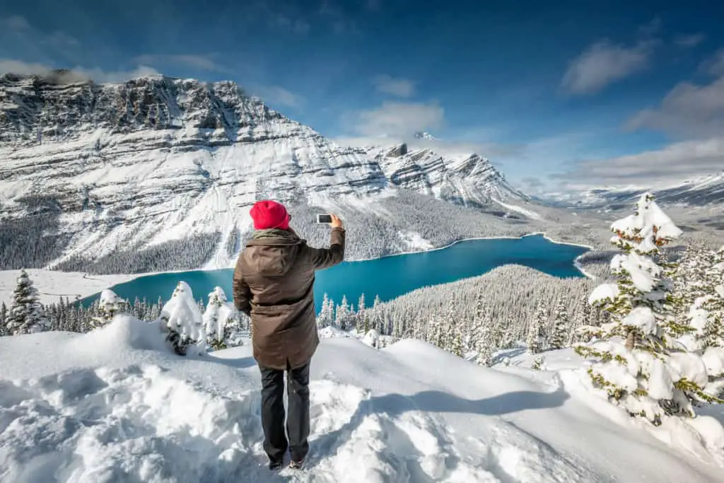 A woman with a red hat is holding a smartphone to take a picture of a snowed in Peyto Lake in October, its water still not frozen