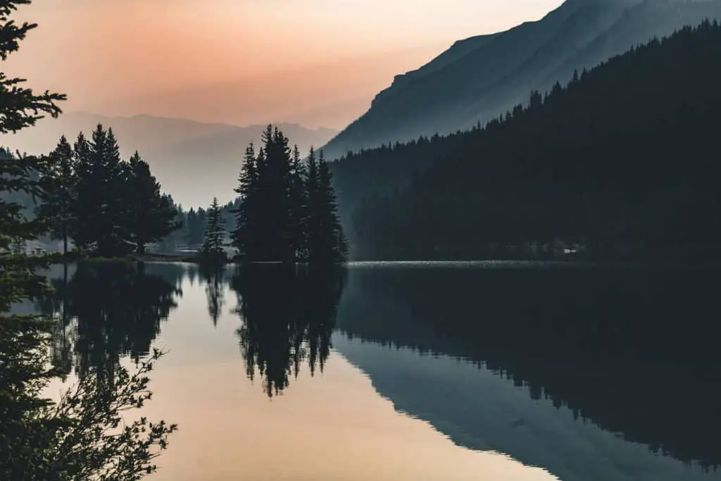 The calm water of Two Jack Lake with cedar trees reflection during an ominous looking sunrise