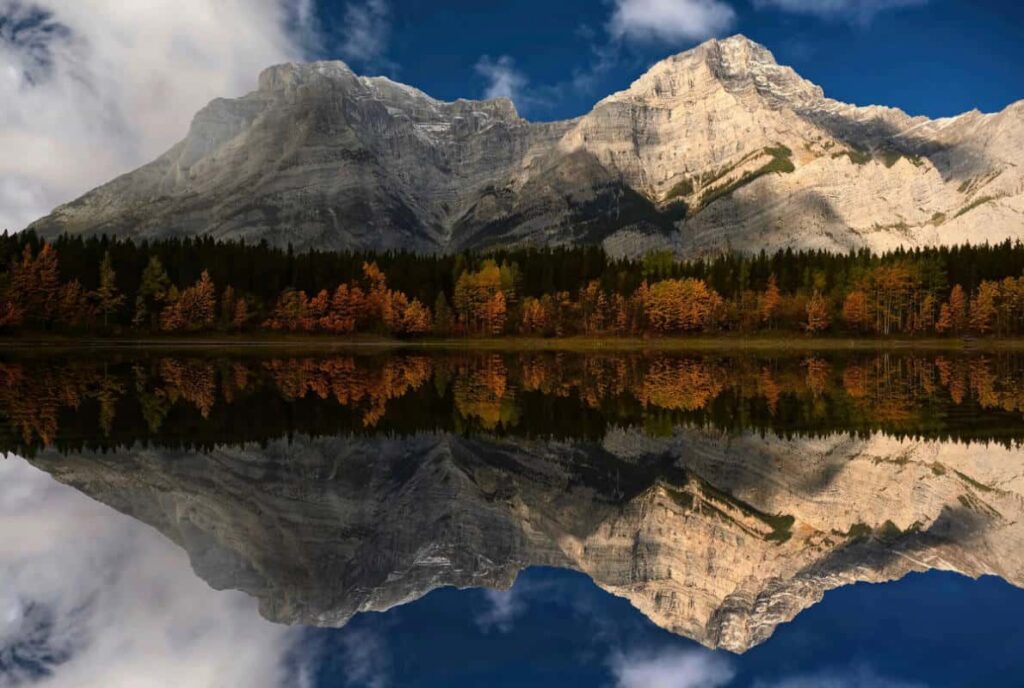 The fall colors of aspen trees surround Wedge Pond in Kananaskis Country, mountains reflect in the still waters