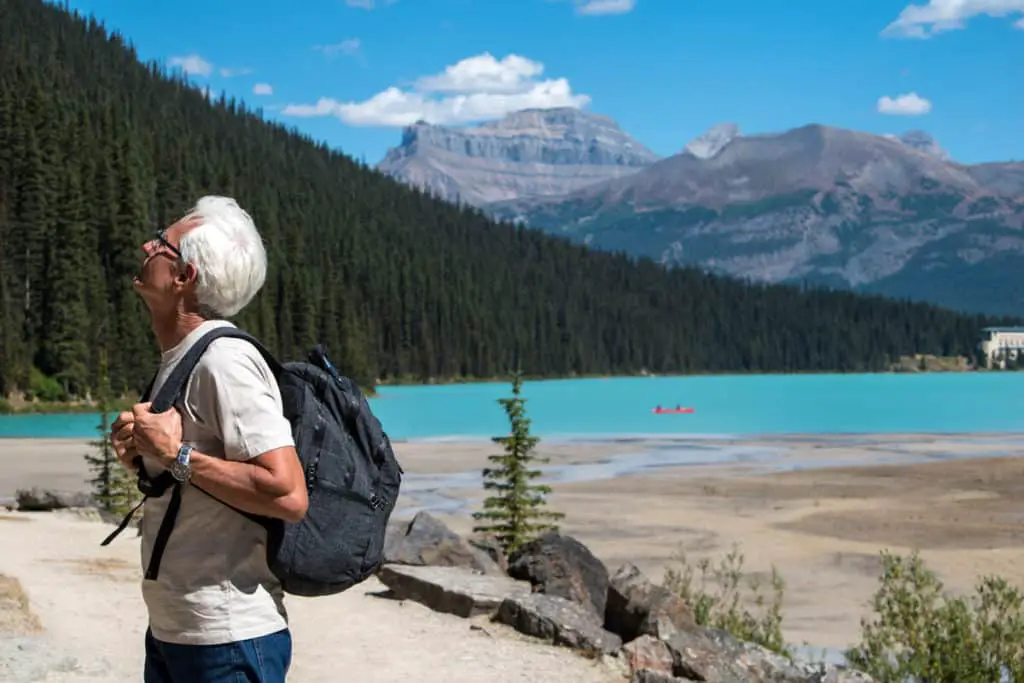 A senior man with white hair looking up a mountain at the other end of Lake Louise, with the lake's turquoise waters in the background