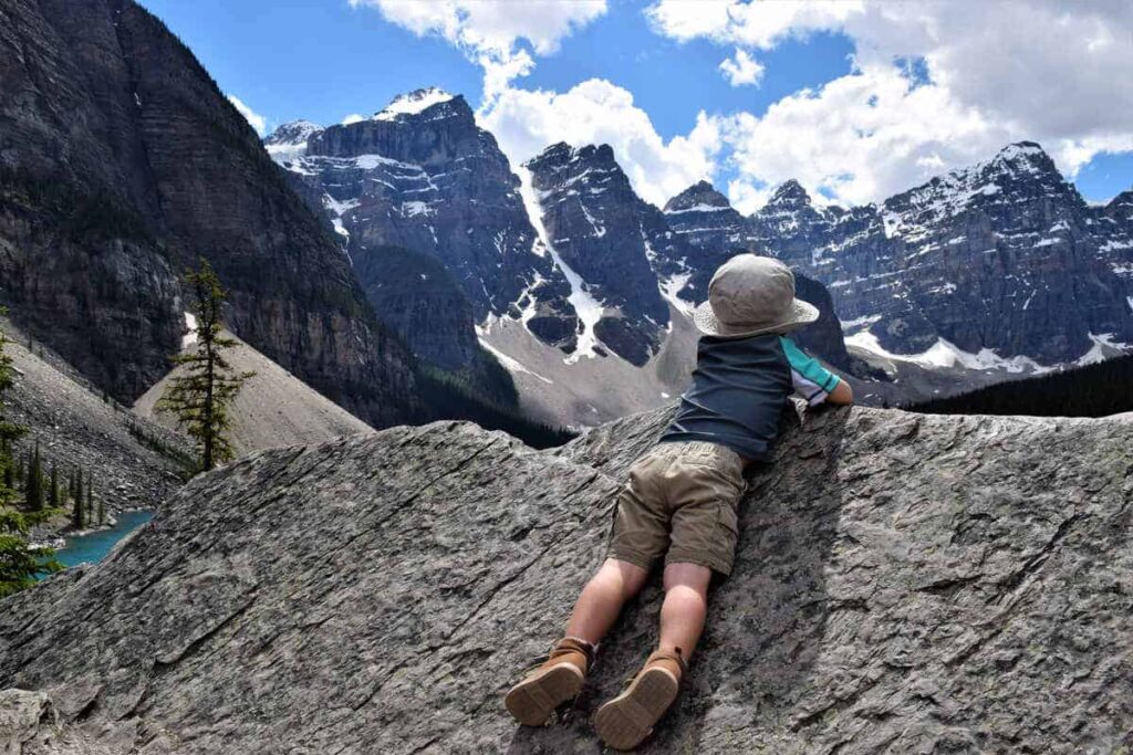 A toddler boy wearing a hat lies on a rock while overlooking Moraine Lake and the Valley of the Ten Peaks